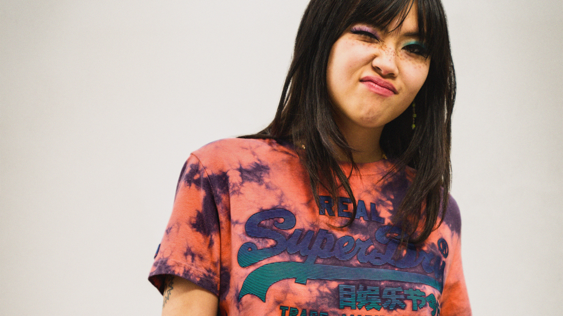An image of a young Asian woman pulling a face with a screwed expression, wearing a bright orange Superdry t-shirt