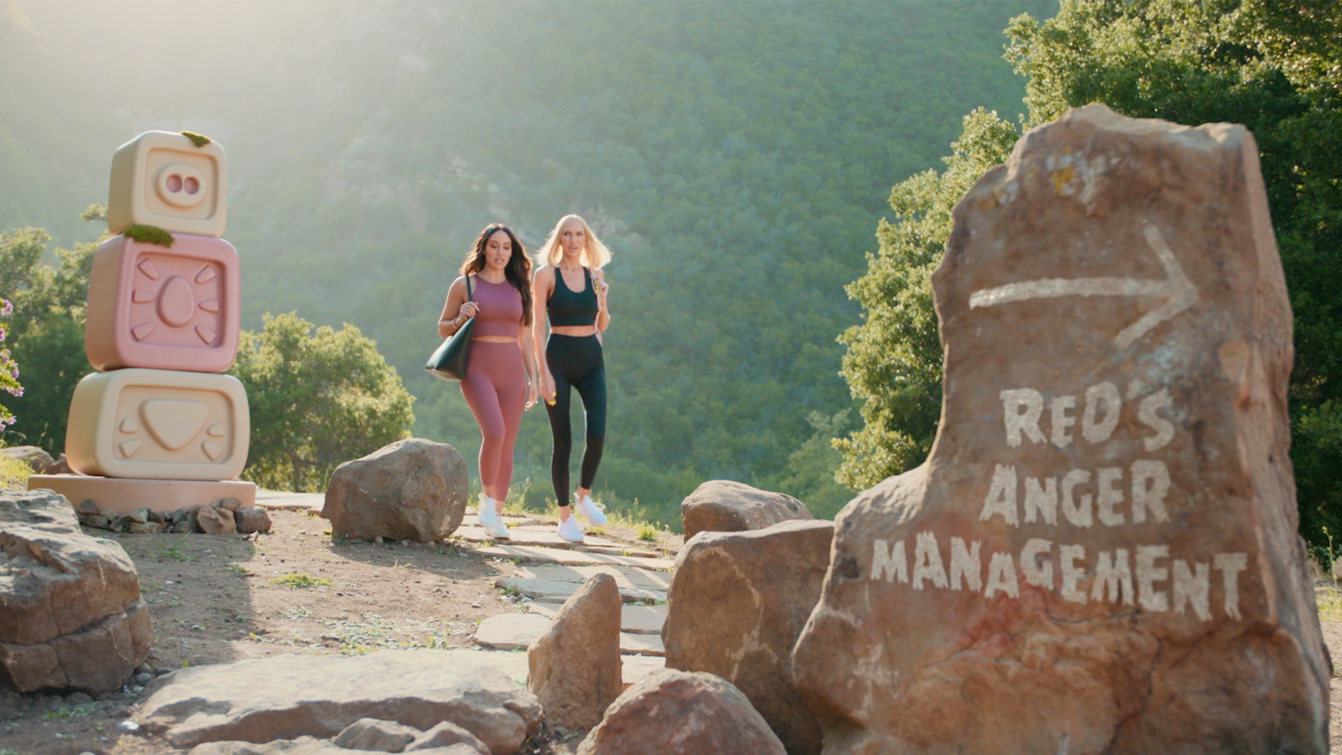 Melissa Gorga and Christine Quinn walking up an outside trail with a sign painted in white on a rock stating Red’s Anger Management with a white arrow.