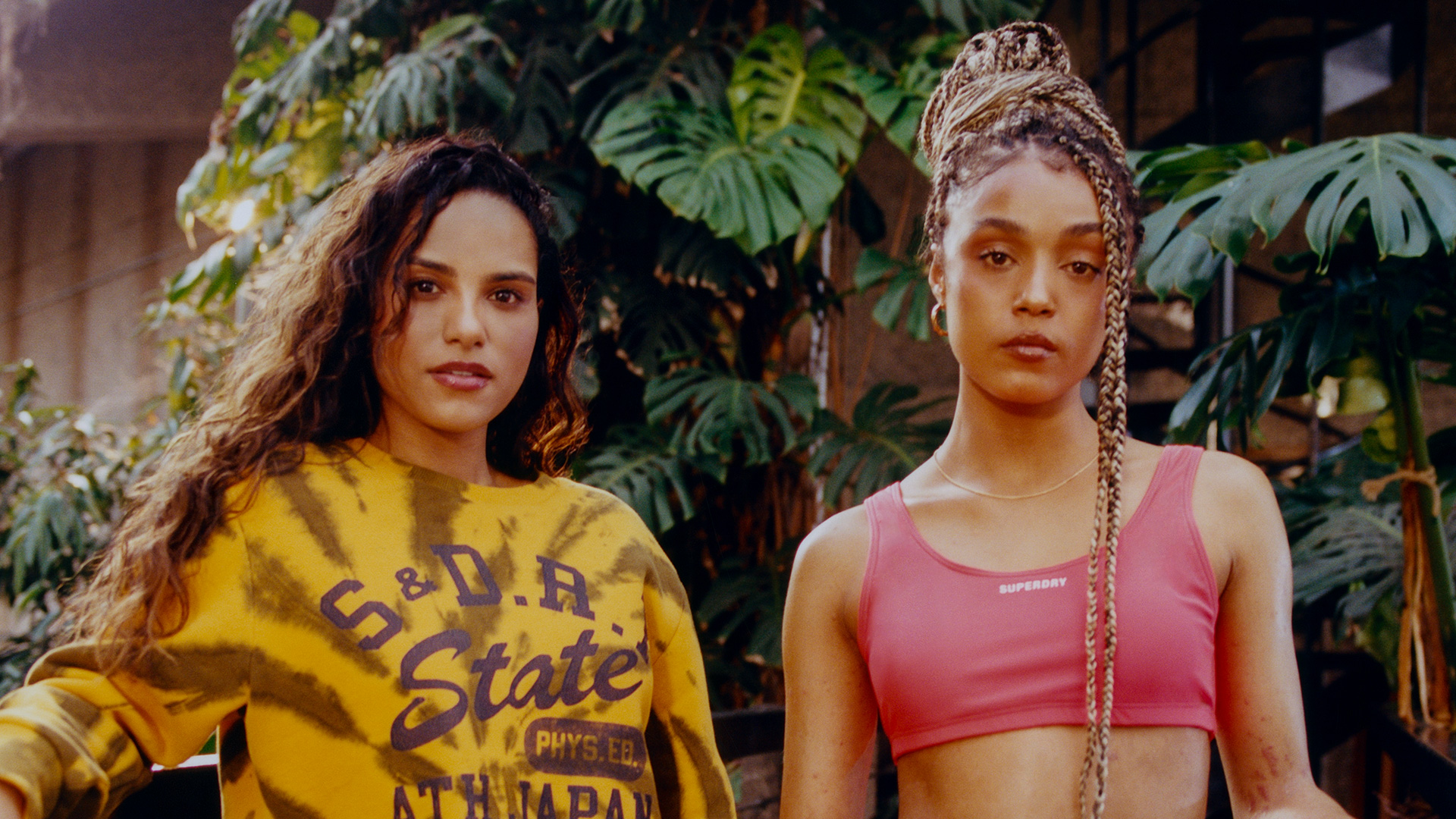 Two young women standing in front of large fronds, the one on the left is wearing a yellow S&DR state jumper and the one on the right is wearing a pink crop top with Superdry in small white lettering
