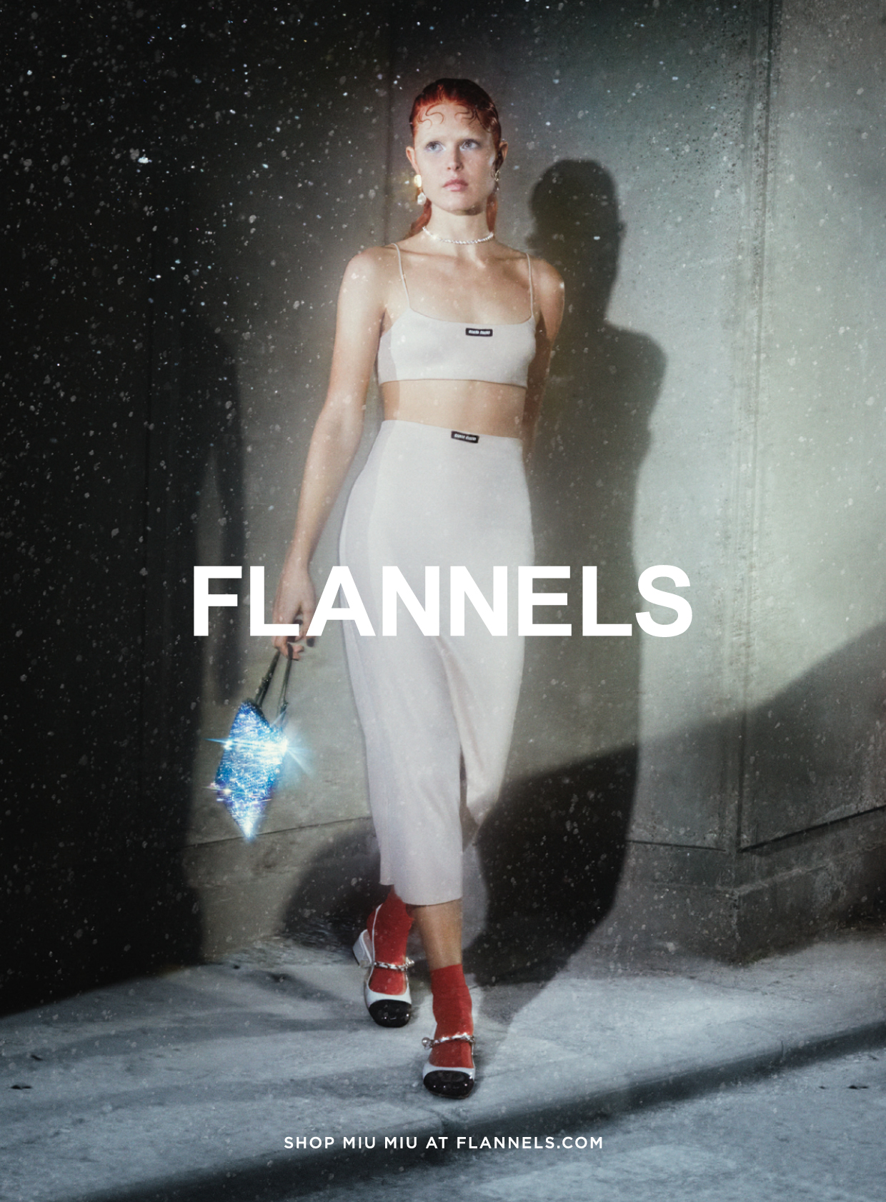 Model in cream top and trousers with red socks and white shoes against a grey concrete background with Flannels logo across the page.